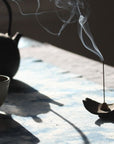 Incense stick with smoke on a textured surface next to a teapot and cup