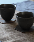 Two black ceramic cups on square coasters on a textured surface.
