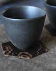 Two dark ceramic cups on a textured surface with a coaster.