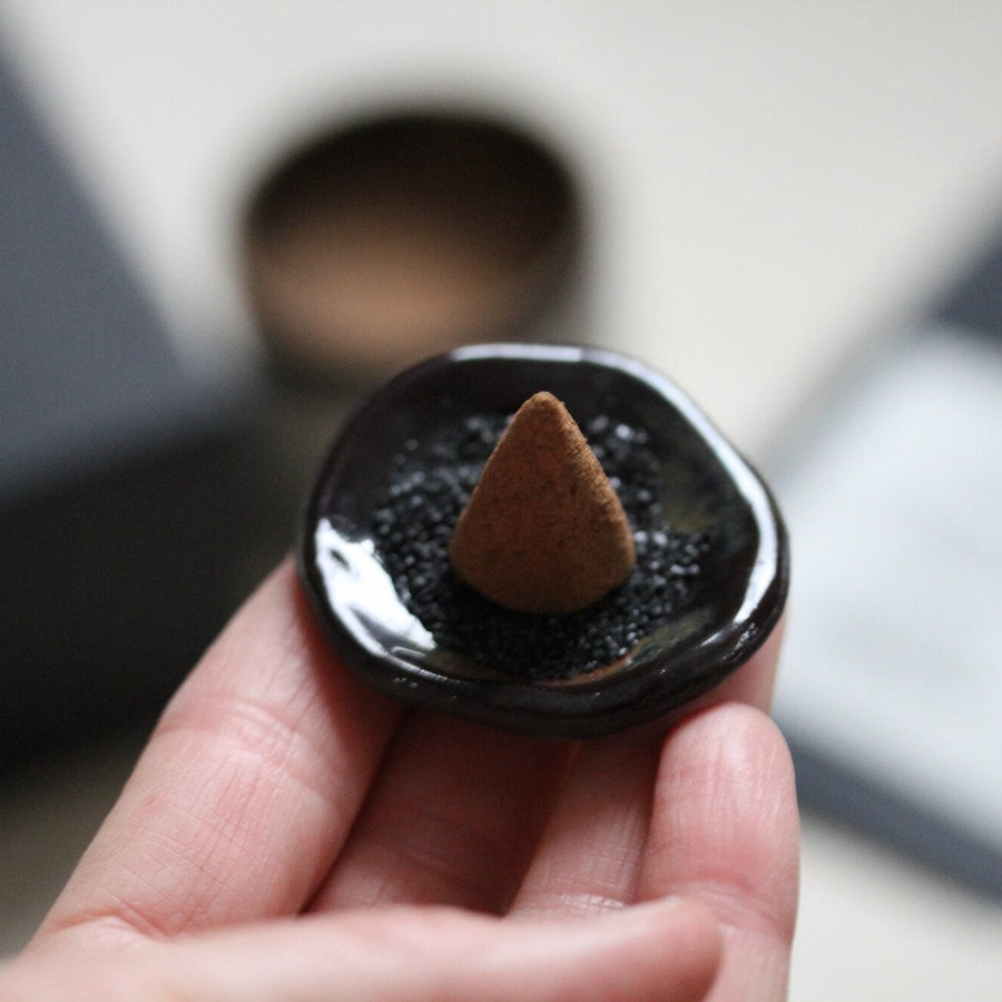 Hand holding a black incense holder with a cone inside, blurred background